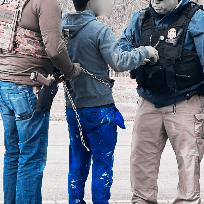 On the left, a man wearing jeans, a brown shirt, black hat, and camo vest tightens chain shackles placed on a man in ripped jeans and a gray hoodie. To his right, a heavy-set man in khakis and a black police vest with a hat appears to be speaking to him. The words say "Ohio man detained in shackles."