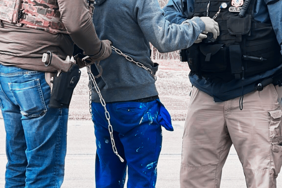 On the left, a man wearing jeans, a brown shirt, black hat, and camo vest tightens chain shackles placed on a man in ripped jeans and a gray hoodie. To his right, a heavy-set man in khakis and a black police vest with a hat appears to be speaking to him. The words say "Ohio man detained in shackles."