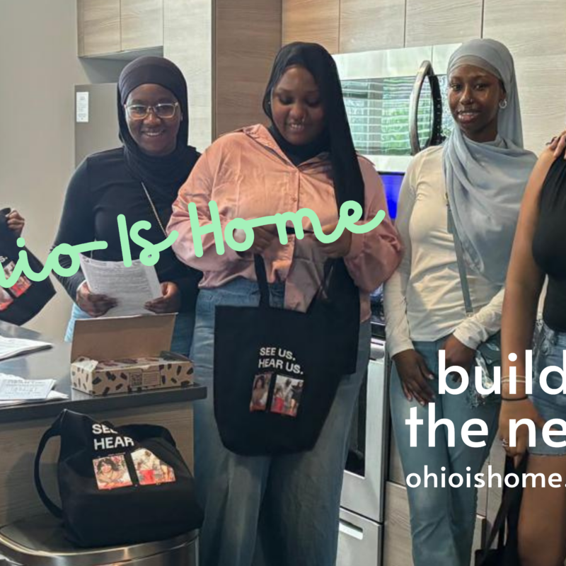 A photo of women and a child, wearing colors of pink, white, black, and blue, holding black tote bags and smiling at the camera. Some of the women have long hair and others are wearing head coverings. The stand in a kitchen next to a trash can and open box. The words read "Ohio Is Home" in navy blue cursive and "build the nest - ohioishome.org" in white plain text.