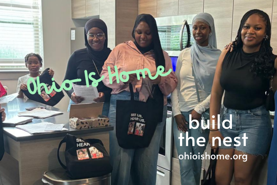 A photo of women and a child, wearing colors of pink, white, black, and blue, holding black tote bags and smiling at the camera. Some of the women have long hair and others are wearing head coverings. The stand in a kitchen next to a trash can and open box. The words read "Ohio Is Home" in navy blue cursive and "build the nest - ohioishome.org" in white plain text.