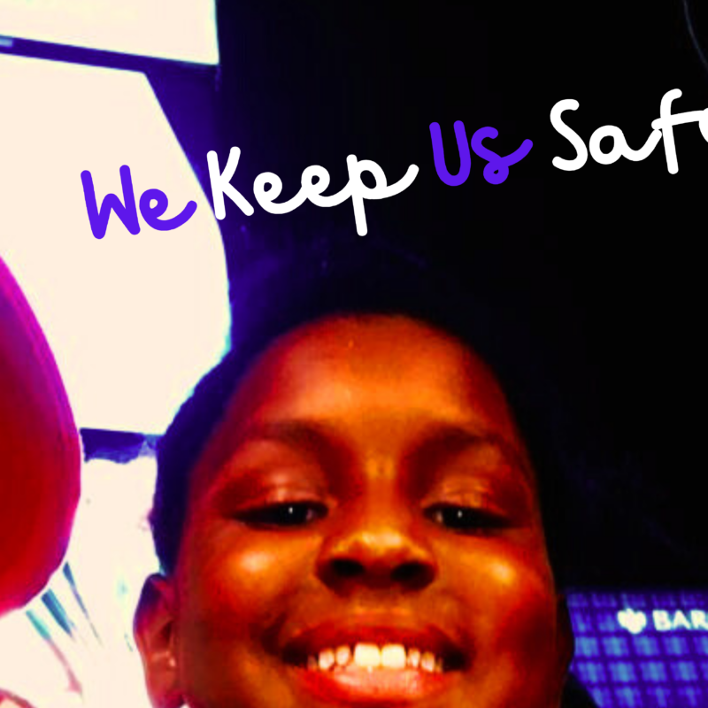 A Black boy wearing a red baseball hat is on the left, with another Black young boy smiling in the middle and a Black man wearing a green striped shirt looks on from the right. Two AAPI individuals appear in the background. The words say, in cursive, "We Keep Us Safe."