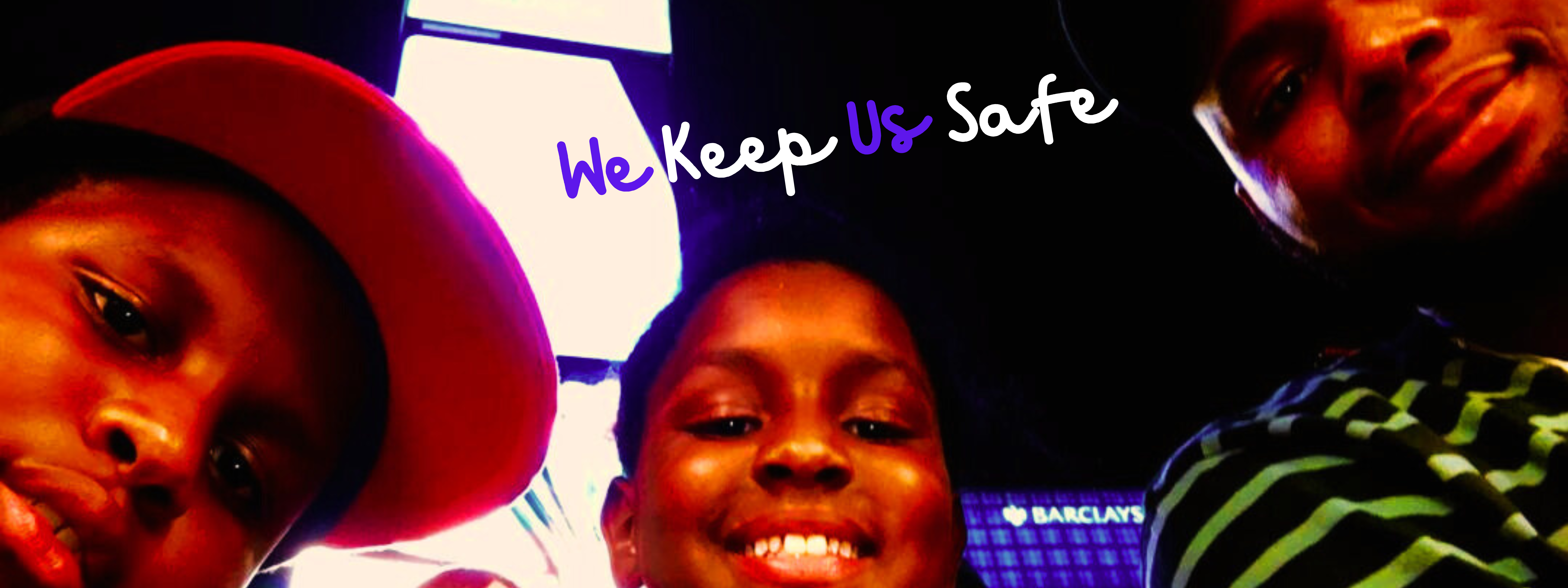 A Black boy wearing a red baseball hat is on the left, with another Black young boy smiling in the middle and a Black man wearing a green striped shirt looks on from the right. Two AAPI individuals appear in the background. The words say, in cursive, "We Keep Us Safe."