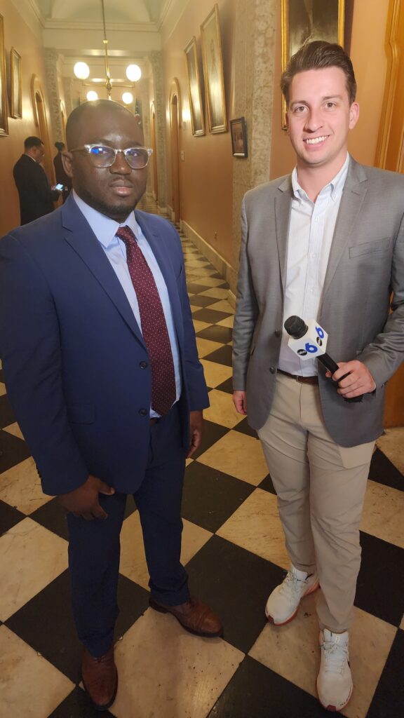 A Black man in a blue suit and red tie stands next to a white man in a gray jacket holding a microphone.