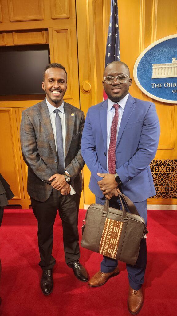 A Black man in a dark gray suit smiles at the camera while standing next to a Black man in a blue jacket and red tie who is holding a briefcase with tapestry details.