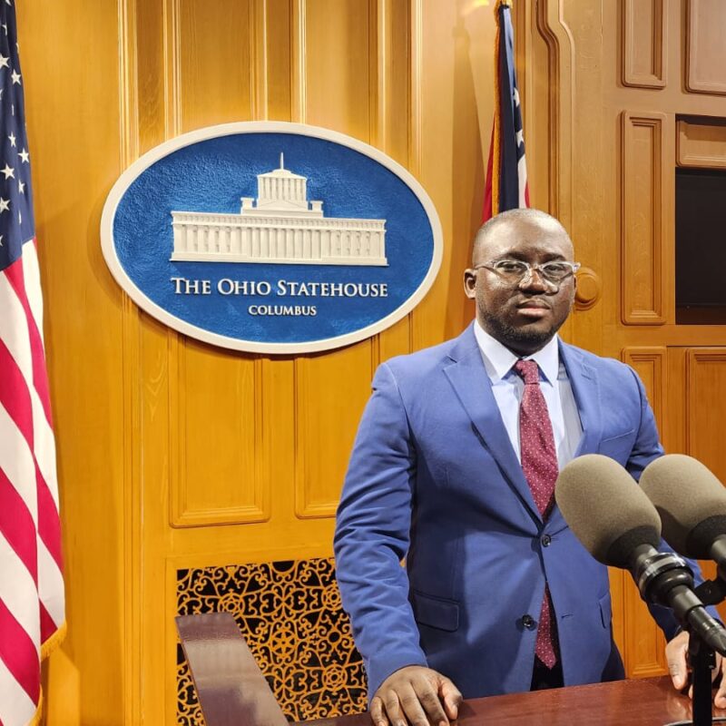 An American flag stands to the left of the Ohio statehouse sign. A Black man with a blue jacket and red tie stands in front of microphones at a podium.