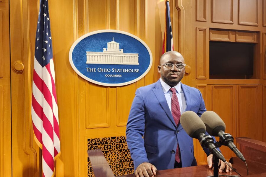 An American flag stands to the left of the Ohio statehouse sign. A Black man with a blue jacket and red tie stands in front of microphones at a podium.