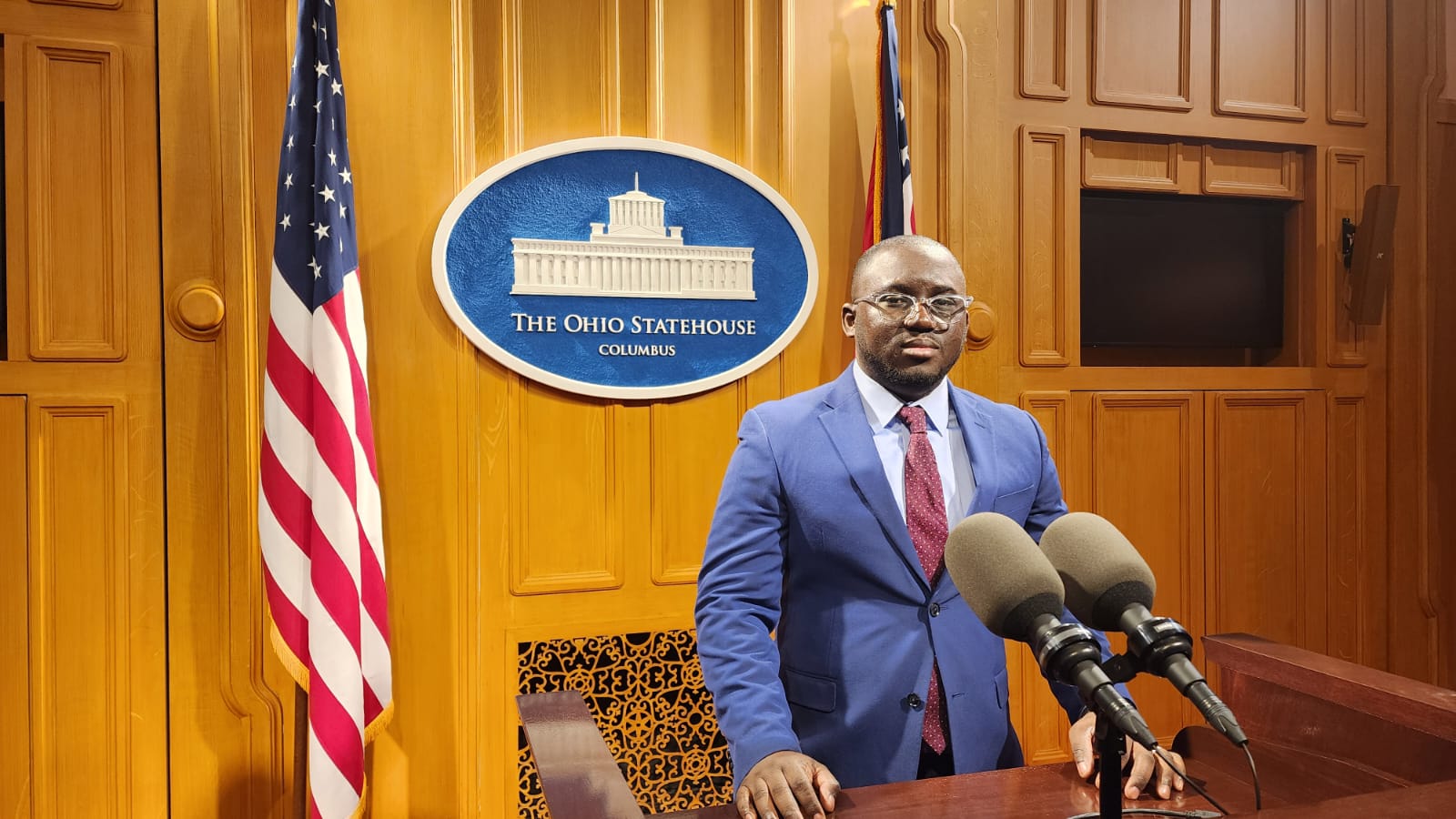 An American flag stands to the left of the Ohio statehouse sign. A Black man with a blue jacket and red tie stands in front of microphones at a podium.