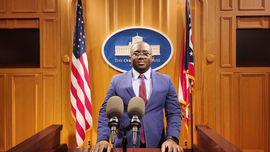 A Black man with a blue jacket and red tie stands in front of microphones at a podium, flanked by two American flags.