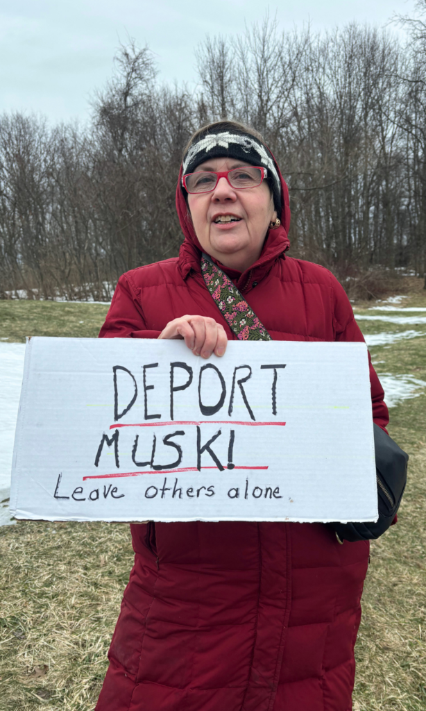 A woman in a red coat holds a sign saying "Deport Musk"