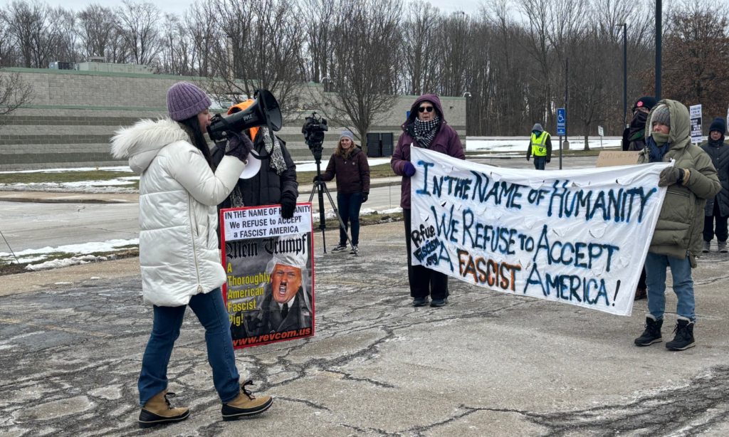 A woman in a cream puffer coat addresses a crowd holding a large banner