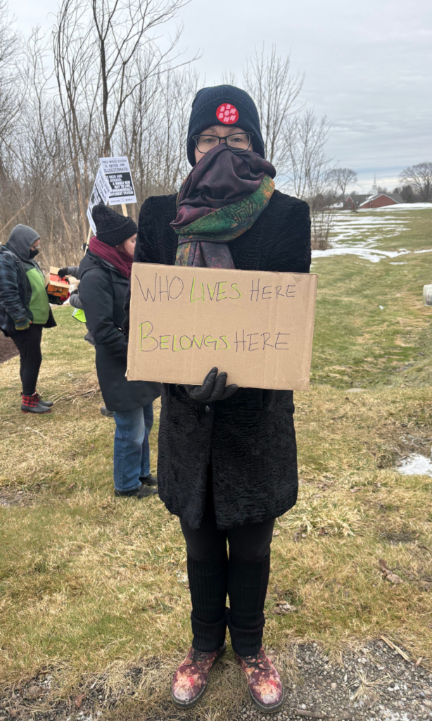 A woman in a mask holds a sign saying "Who Lives Here, Belongs Here"