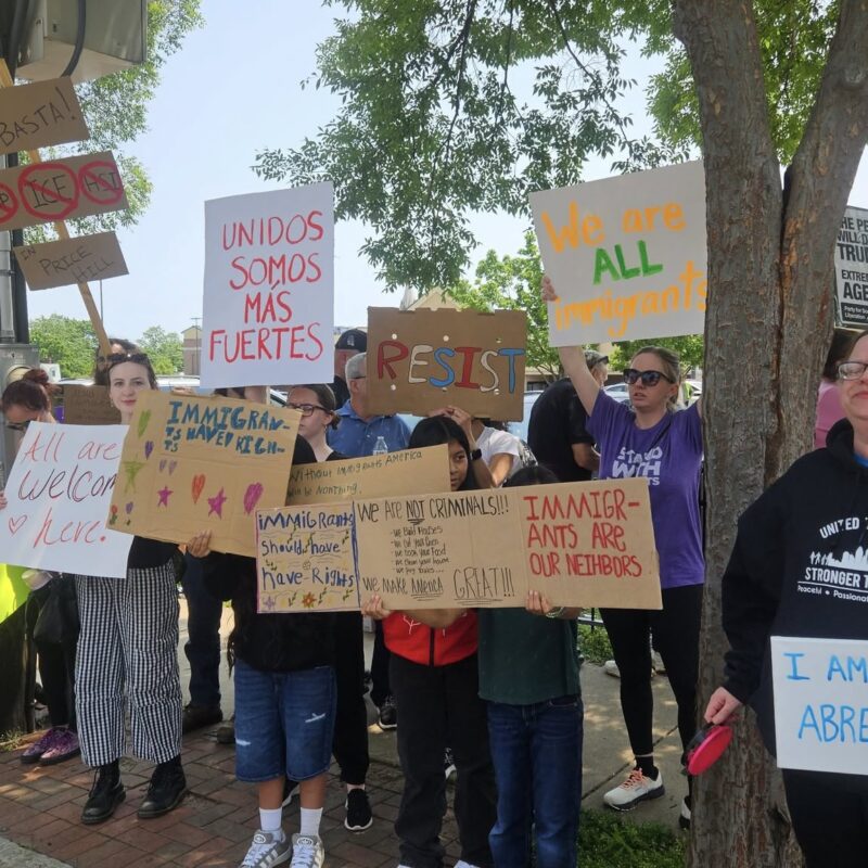 A group of people line a sidewalk carrying various signs in different colors expressing support for immigrants' rights. There are trees in the background.