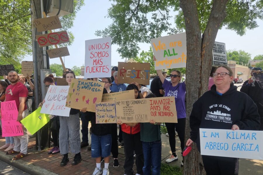 A group of people line a sidewalk carrying various signs in different colors expressing support for immigrants' rights. There are trees in the background.